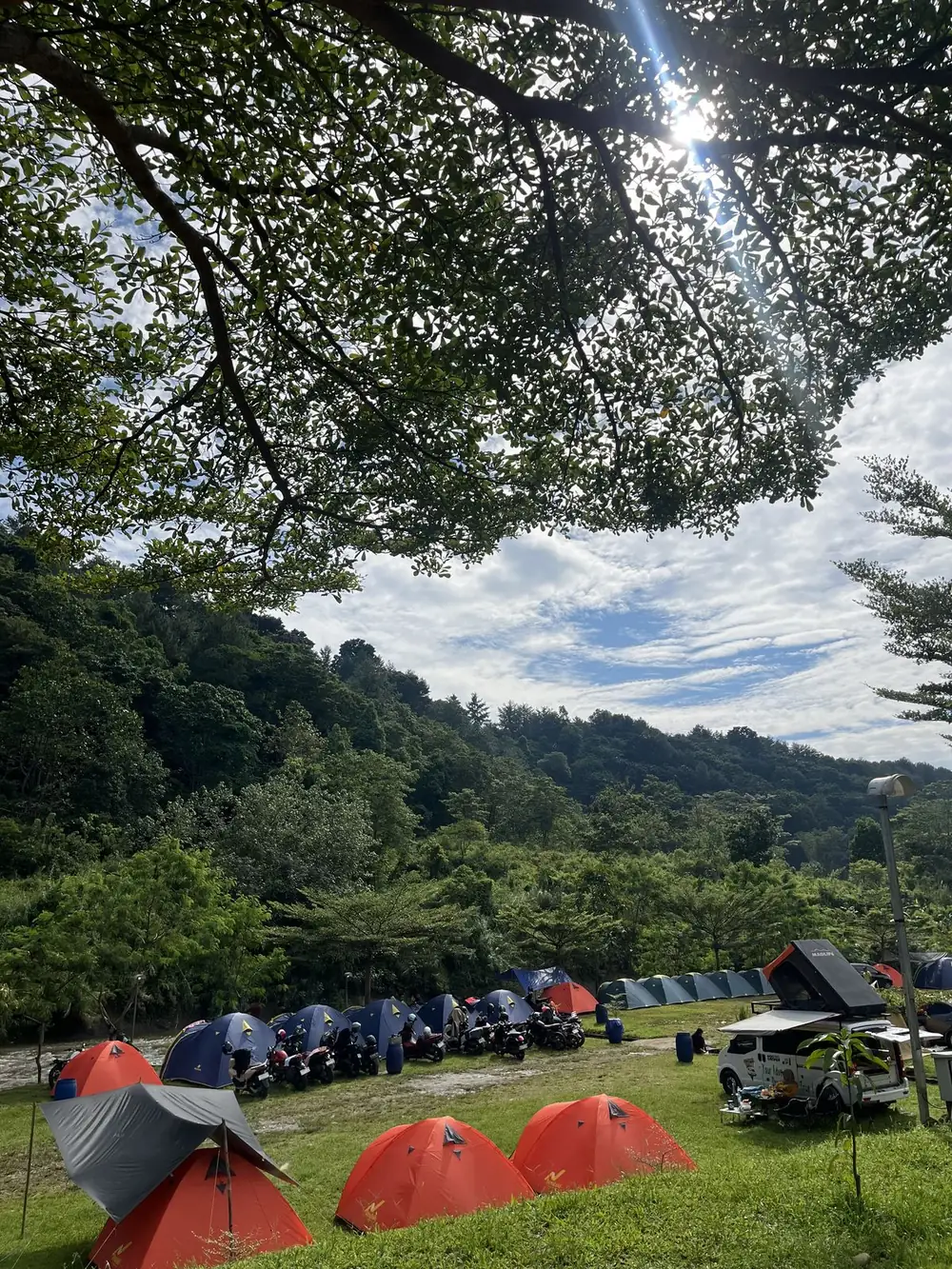 Tenda-tenda di padang rumput Sisi Cai di bawah rindang pohon dengan perbukitan hijau